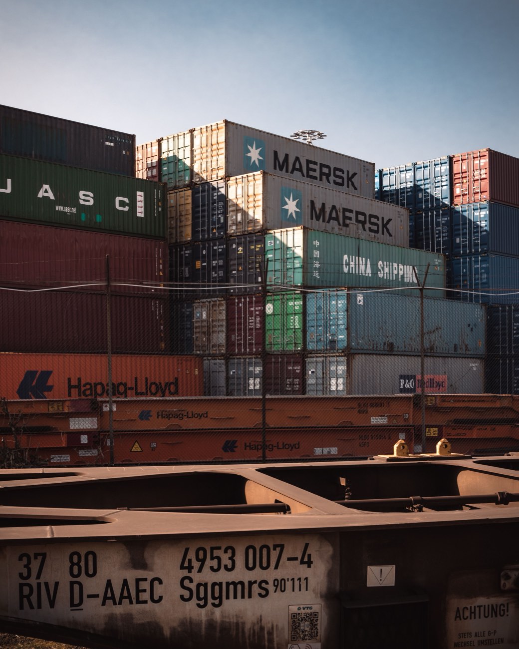 Stacked intermodal containers behind a fence, with a well car in the foreground